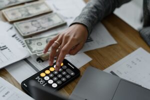 Person using a calculator with stacks of cash and papers next to them on a table