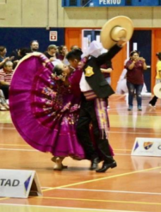 a couple dancing at a competition; the man wears a dark suit and lifts a straw hat dramatically in the air while a woman in a full, maroon dress clings to him, her skirt billowing as they move
