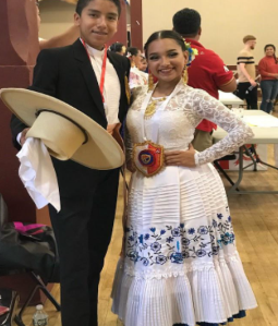 a young couple wearing formal dance attire stand together, smiling at the camera. The young man gestures with his wide-brimmed hat while the young woman in white lace rests a hand on her large, colorful belt