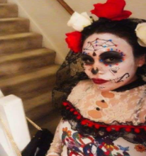 the writer wears a colorful costume -- white-painted face with dark eyes and nose suggesting a skull, a colorful spider web on the forehead and large white and red roses pinning a black lace veil to her head
