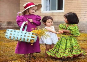 photo shows three young children representing the different peoples of Chihuahua: a girl in a long-sleeved pink dress and straw bonnet carrying a basket; a toddler wearing white; and a dark-haired girl in a colorful green patterned dress with a flounced skirt