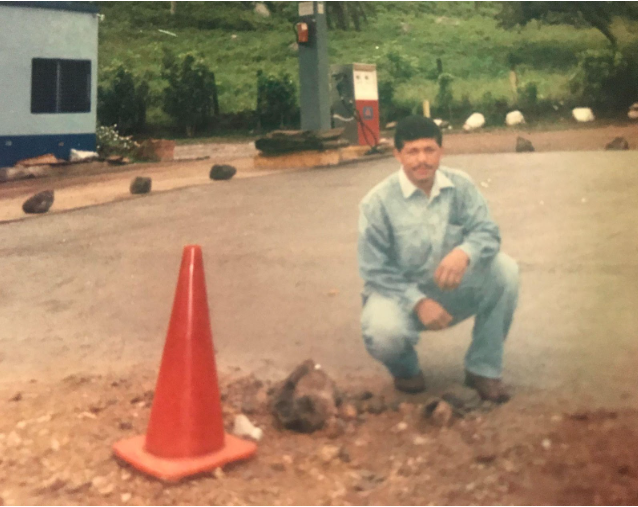 faded photograph shows a dark-haired man with a mustache, wearing denim, crouched in the dusty foreground near a traffic cone, a gasoline pump in the background