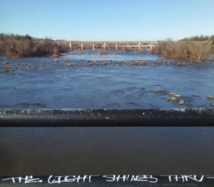 photograph of James River, rocky and blue, with a distance arched bridge in the background and, in the foreground, a bridge railing with the graffiti words "THE LIGHT SHINES THRU"