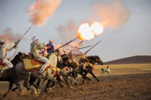 a photo shows a group of men on horseback, racing across a desert landscape and firing long rifles that burst with fire and smoke