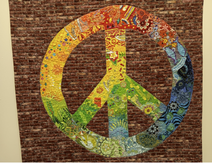 photograph of a very colorful quilt wall hanging. A large peace sign stands out against a background of fabric with a pattern of red bricks. Bright yellows, greens, oranges, reds, and blue fabrics with vivid patterns make up the peace sign.