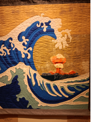photograph of a colorful quilt wall hanging, showing a large blue wave in the style of a Japanese woodblock print. In the sunset-colored background, an intense orange mushroom cloud is visible.
