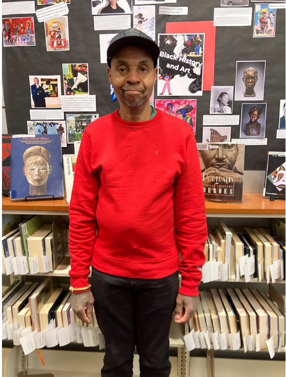 photograph of the author, Lloyd, wearing a read sweatshirt and a baseball cap, standing in front of a display of books, looking frankly into the camera