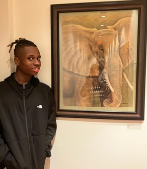 photo of author, smiling and wearing a black hoodie, standing in front of a large framed painting of an elephant. The elephant appears to be looming, it's large head -- big ears, tusks, curled trunk -- more than filling the frame