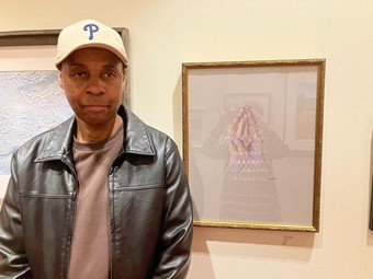 photo of the author, wearing a baseball cap and leather jacket, turning to look at a framed painting painting showing two ornate houses. In front of them stretches a grey street with walking figures.