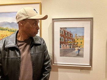 photo of author, wearing a leather jacket and baseball cap, standing in front of a framed drawing of two praying hands