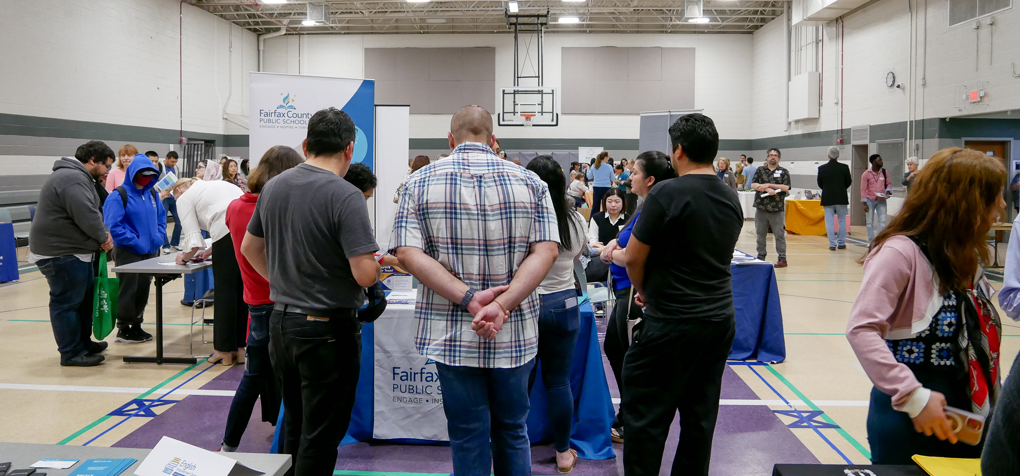 Diverse adults mingle at a busy career fair, visiting tables from multiple employers such as Fairfax County Public Schools