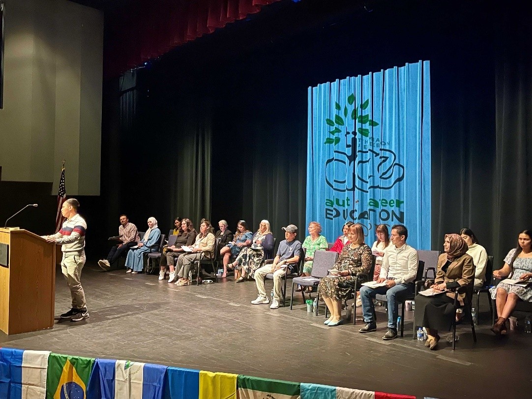 photograph shows adult learners lined up across a stage, preparing for their turn at the podium
