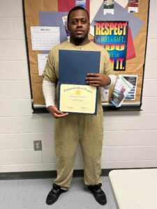 A young man wearing an institutional uniform stands in front of a colorful bulletin board decorated with a poster reading RESPECT. He smiles as he holds up a mounted, gold-sealed certificate.