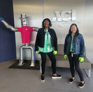 photograph two women stand in an office hallway in front of the ACI logo; a large silver robot, wearing a red T-shirt and waving is nearby