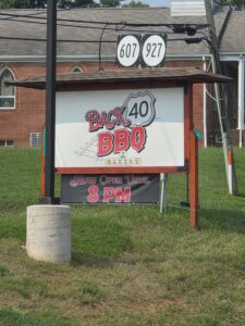 on a grassy roadside is Back 40 BBQ's large, wooden, billboard-style sign. Beneath the bold restaurant logo is a banner proclaiming Open Until 8 PM