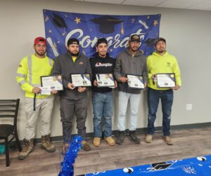 A group of five young men, some wearing construction workers' neon yellow, hold certificates in front of a large "Congrats!" banner