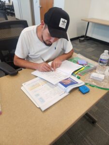 a young man in a baseball cap studies at a table, notebook and textbook open in front of him
